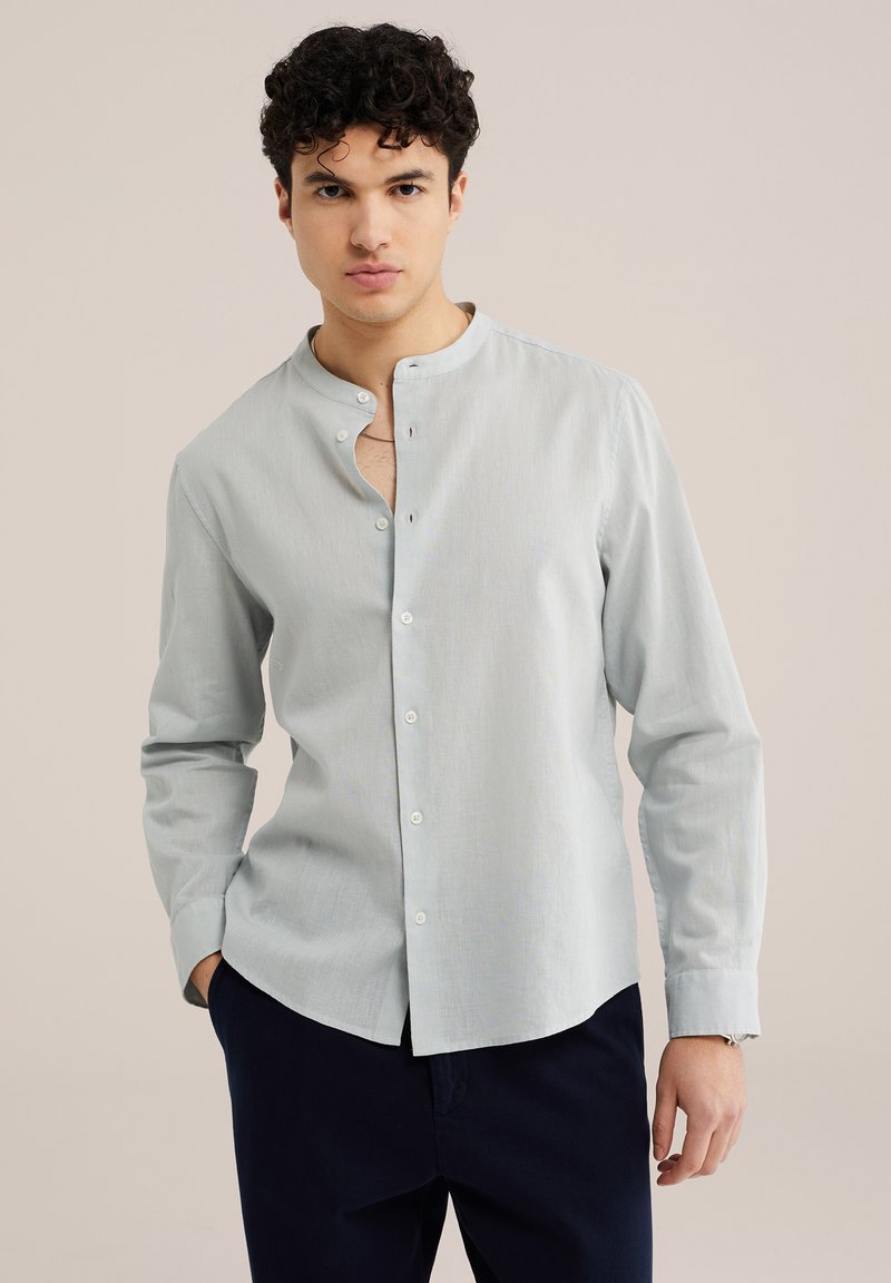 Jeune homme aux cheveux bouclés portant une chemise grise clair à col mandarin et un pantalon foncé, debout devant un fond uni.