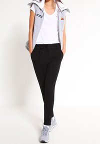 Haut sans manches gris avec logo, t-shirt blanc, pantalons de jogging noirs et baskets grises. La tenue présente un style décontracté et athlétique.