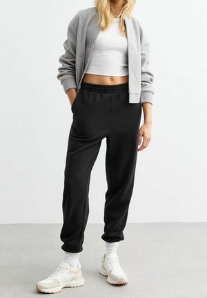 Kobieta ubrana w szarą kurtkę bomber, biały krótki top, czarne spodnie dresowe, białe skarpetki oraz beżowo-białe sneakersy, stojąca przy białej ścianie.