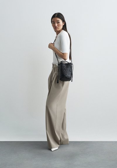 Μαύρη τσάντα τύπου bucket από πλεκτό υλικό με κλείσιμο τύπου drawstring. Υφή επιφάνειας και δύο χερούλια. Το μοντέλο φοράει λευκό ριγέ μπλουζάκι και μπεζ παντελόνι σε φαρδιά γραμμή.