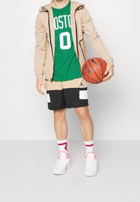 Béžová mikina na zip, zelené tričko s nápisem "BOSTON", béžovo-černé basketbalové šortky s bílými panely, bílé tenisky s výraznými prvky, drží basketbalový míč.