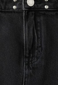 Gros plan sur la ceinture d'un jean en denim noir avec bouton métallique et rivets, coutures visibles et passants de ceinture.