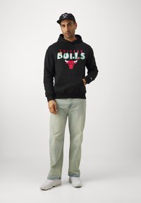 Felpa nera con grafica "Chicago Bulls" e logo del toro, abbinata a jeans azzurri chiari e sneakers bianche, indossata con un cappellino nero.