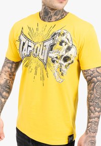 Geltonas medvilnės marškinėliai su grafika, kurioje pavaizduotas kaukolė ir tekstas "TAPOUT", akcentuotas juodais ir pilkais sprogimais.