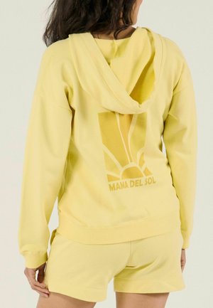 Vrouw die een gele hoodie draagt met een zonafbeelding en de tekst "MAMA DEL SOL" op de achterkant, gecombineerd met bijpassende gele shorts.