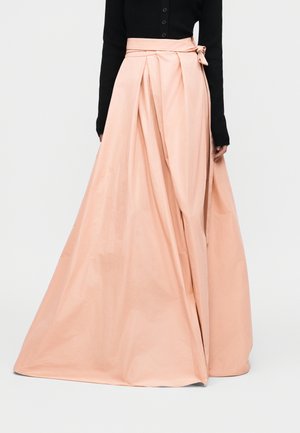 Μακριά πτυχωτή φούστα σε ροδακινί απόχρωση με φιόγκο στη μέση στο πλάι, φορεμένη με μαύρο μπλουζάκι μακρυμάνικο.