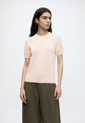Femme aux longs cheveux foncés portant un haut rose clair à manches courtes et un pantalon ample vert olive, debout devant un fond clair uni.