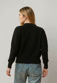 Schwarzer gerippter Strickpullover mit rundem Halsausschnitt, langen Ärmeln und taillierter Passform. Kombiniert mit hellen Jeans.