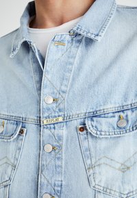 Chaqueta de mezclilla azul clara con cierre de botones, costuras amarillas y dos bolsillos en el pecho. Presenta una etiqueta metálica "REPLAY" y botones plateados.