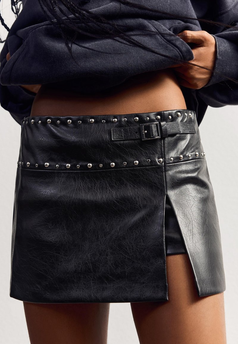 Mini-jupe noire en simili cuir avec des clous argentés, une ceinture à boucle et une fente latérale avant, portée avec un haut sombre légèrement remonté pour dévoiler la taille.