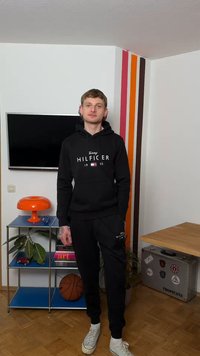 Young man wearing black Tommy Hilfiger tracksuit stands indoors near a shelf with books, basketball, orange lamp, and striped wall decor.