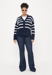 Ναυτικό πουλόβερ με λευκές ρίγες, V-λαιμόκοψη, κλείσιμο με κουμπιά, ραβδωτή υφή, σε συνδυασμό με σκούρες φαρδιές τζιν παντελόνες. Αξεσουάρ περιλαμβάνουν γυαλιά ηλίου.