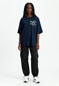 T-shirt ample navy en coton, arborant le texte « Mi Amor » et un design en forme de cœur. Associe à un pantalon cargo noir et des baskets blanches.