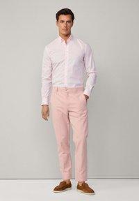 Camicia button-up rosa chiaro, slim-fit con righe, abbinata a pantaloni rosa chiaro e mocassini in suede marroni, in piedi contro uno sfondo neutro.