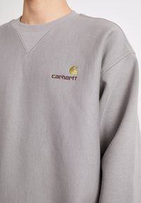 Sudadera gris de algodón con cuello redondo, que presenta un logo "carhartt" marrón y un bordado de icono amarillo en el pecho.