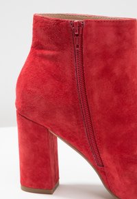 Bottine en daim rouge avec un talon épais et un détail de fermeture éclair latérale. Texture lisse, couleur profonde et design minimaliste.