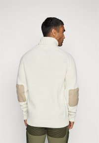 Maglione lavorato a maglia color crema con colletto alto e toppe beige sui gomiti, caratterizzato da polsini e orlo a coste, visto da dietro.