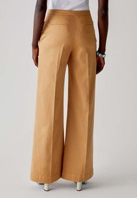 Pantaloni a gamba larga di un caldo color tan, realizzati in tessuto morbido. Presentano tasche posteriori e una cintura su misura per una vestibilità pulita.