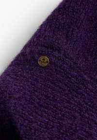 Tela de punto morada con una textura suave, detallada con un botón de bronce que presenta un diseño de calavera. Vista cercana que destaca las costuras.