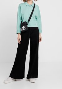 Osoba nosząca miętowo-zielony top z długim rękawem, szerokie czarne spodnie, białe sneakersy i małą czarną torebkę typu crossbody Adidas.