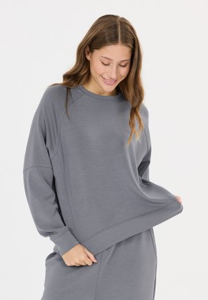 Jonge vrouw glimlacht terwijl ze een loszittend grijs shirt met lange mouwen draagt en er zachtjes aan trekt tegen een lichte achtergrond.