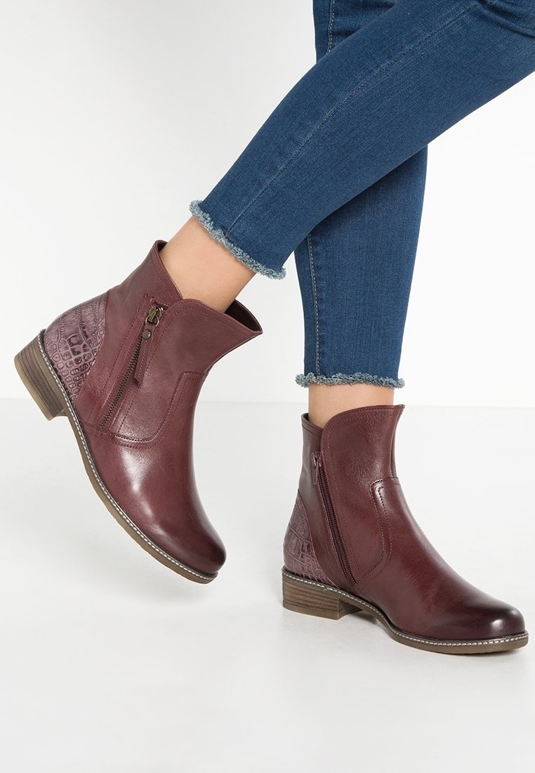 Bottes en cuir bordeaux montant à la cheville avec des fermetures éclair latérales, panneau arrière texturé, portées avec un jean skinny bleu à ourlet effiloché.
