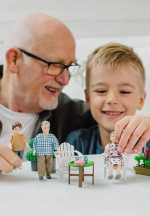 Neljä värikästä muovihahmoa on aseteltu vihreälle pöydälle, jossa on vaaleanpunainen lautanen. Kaksi valkoista tuolia ja ruukkukasveja on myös näkyvissä.