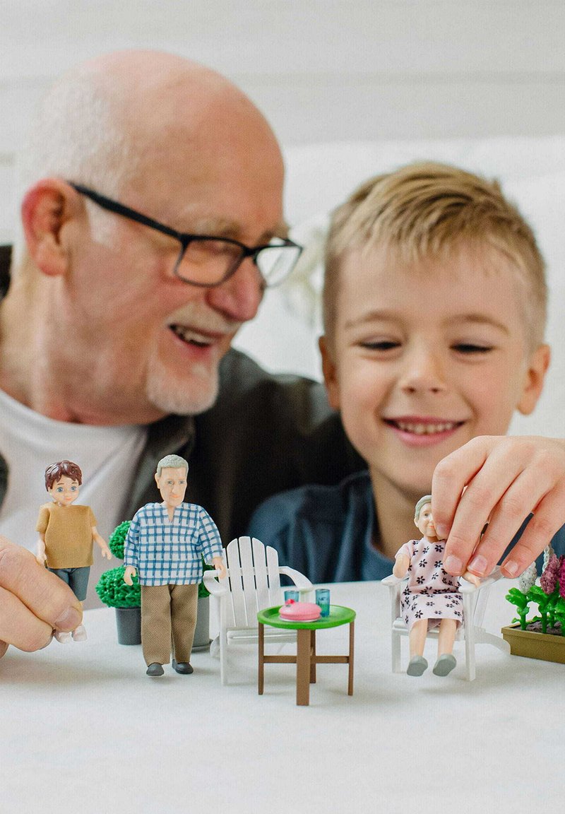 Vier bunte Plastikfiguren sind an einem grünen Tisch mit einem rosa Teller angeordnet. Zwei weiße Stühle und Topfpflanzen sind ebenfalls sichtbar.