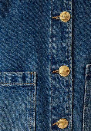 Denimjack-detail met blauwe stof, gouden knopen en twee voorkanten. Zichtbare stiksels langs de randen, wat textuur toevoegt.