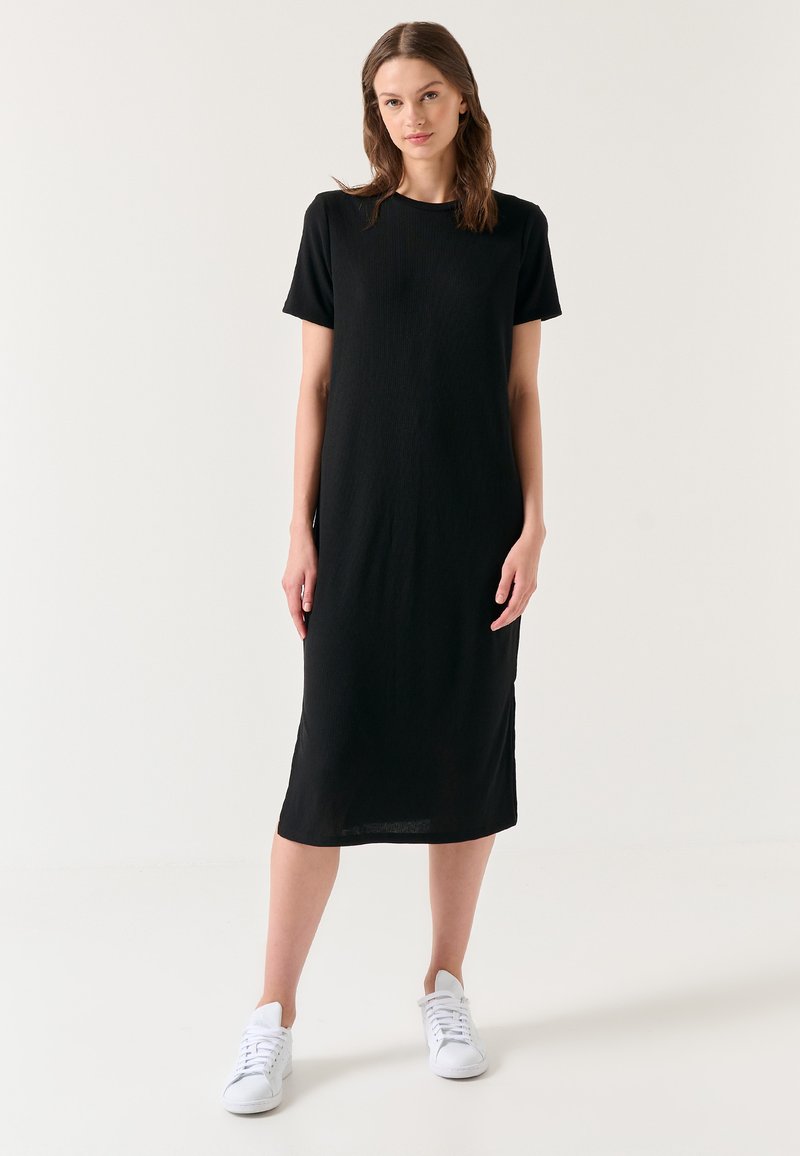 Jeune femme debout portant une robe midi noire unie à manches courtes et des baskets blanches sur fond blanc.