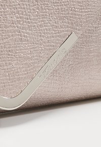 Metallinen vaaleanpunainen käsilaukku, jossa on teksturoitu pinta; siinä on hallitseva hopeinen logo-aksentti ja kulmikas metallinen yksityiskohta reunan varrella.