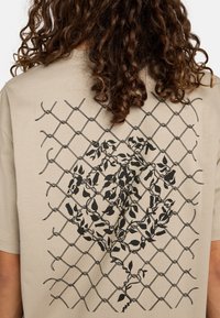 Beige cotton t-shirt featuring a black graphic design of a chain-link fence with floral elements in the center. Short sleeves, crew neck.