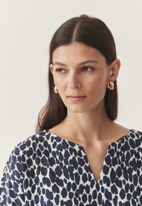 Plava i bijela majica s leopard printom s V-izrezom, izrađena od mekog materijala i opuštenog kroja. Zlate kružne naušnice dopunjuju dizajn.