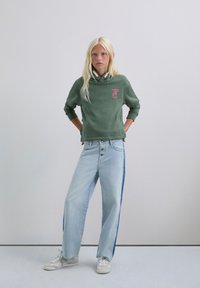 Sudadera verde con un logo sutil, combinada con jeans de pierna ancha azul claro que presentan una franja lateral azul oscura. Zapatillas blancas completan el conjunto.