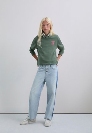 Sudadera verde con un logo sutil, combinada con jeans de pierna ancha azul claro que presentan una franja lateral azul oscura. Zapatillas blancas completan el conjunto.