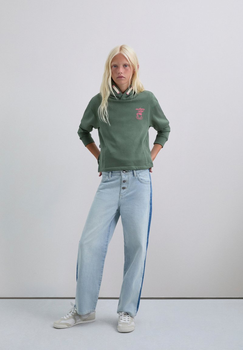 Sudadera verde con un logo sutil, combinada con jeans de pierna ancha azul claro que presentan una franja lateral azul oscura. Zapatillas blancas completan el conjunto.