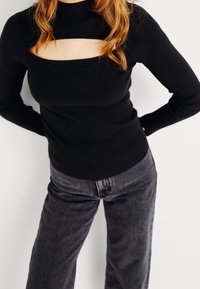 Svart ribbad långärmad topp med hög hals och rektangulär utskärning som har en figurnära silhuett. Parat med mörkgrå jeans.