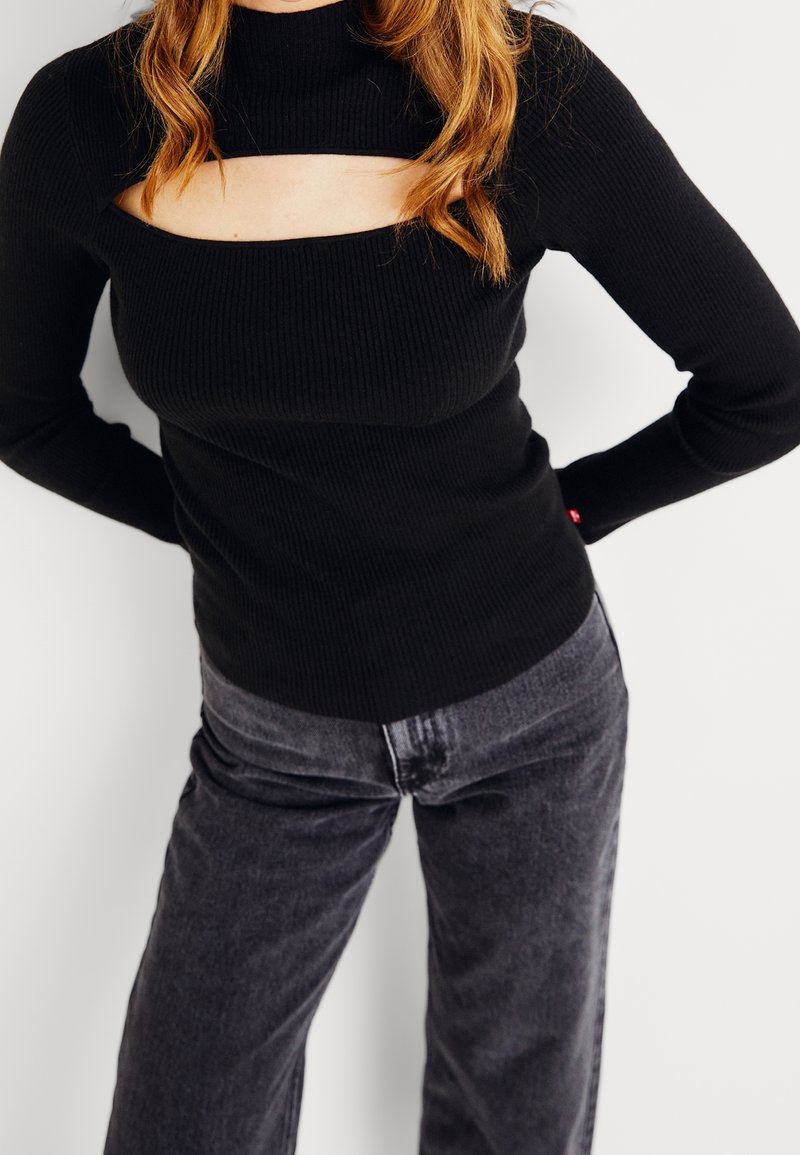 Svart ribbad långärmad topp med hög hals och rektangulär utskärning som har en figurnära silhuett. Parat med mörkgrå jeans.