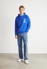 Sudadera azul con detalles gráficos en blanco, bolsillo tipo canguro, puños acanalados, combinada con jeans azul claros y zapatillas blancas sobre un suelo de concreto.