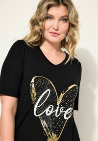 Svart kortärmad V-ringad t-shirt med ett guld- och silverhjärta där ordet "love" är skrivet i vit kursiv stil, dekorerad med rhinestones.