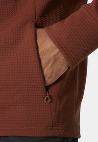 Tela de color óxido con un patrón de cuadrícula texturizada. Presenta un bolsillo lateral con un tirador de cremallera marrón, mostrando un diseño elegante y funcional.