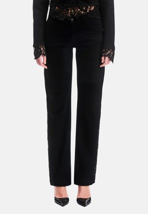 Frau trägt schwarze High-Waist-Hose mit Spitzendetails am Bund, an den Bündchen und an den Seiten, kombiniert mit schwarzen spitzen Schuhen.