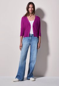 Fuchsia cardigan met driekwartmouwen, knoopsluiting, gecombineerd met lichtblauwe wijdlopende jeans en witte sneakers op een neutrale achtergrond.