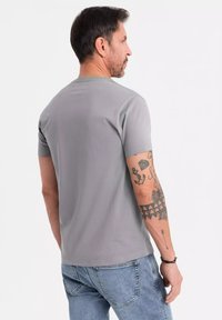 Camiseta de manga corta gris, hecha de algodón suave, con cuello redondo y corte relajado. Tatuajes visibles en el brazo añaden detalle.