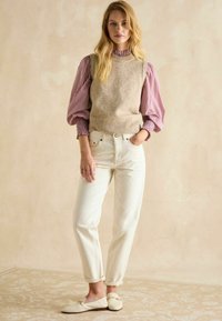 Beige gestrickte Weste über einer rot-weißen gestreiften Bluse, kombiniert mit weißen Jeans in lässiger Passform und hellen Mokassins. Strukturierter Hintergrund.