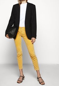 Blazer negro sobre una camiseta blanca, combinado con pantalones de pana amarillos. Bolso negro y sandalias negras con tiras. Diseño simple y moderno.