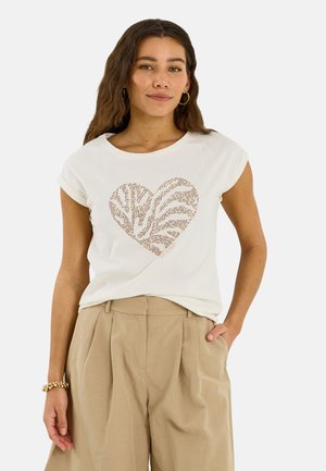 Mujer con cabello largo y castaño lleva una camiseta blanca con un diseño de corazón y pantalones beige de talle alto y plisados, posando con una mano en el bolsillo.