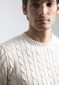 Jersey de punto grueso en color blanco roto, con un amplio cuello redondo y un patrón texturizado con cables en relieve. Tejido suave con un grosor considerable.