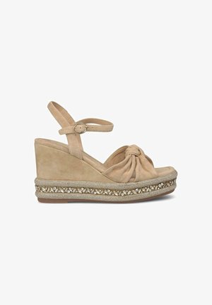 Beige suede wedge sandals with ankle strap, knotted front design, and decorative metallic trim featuring small gemstones along the sole.