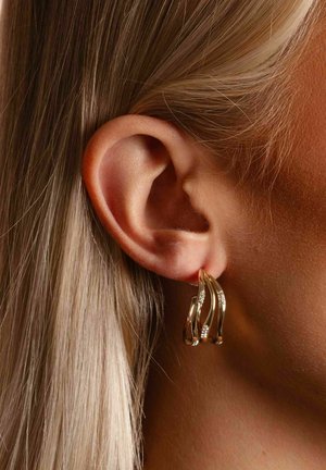 Nahaufnahme eines Frauenohrs mit glattem blondem Haar, geschmückt mit einem goldenen Ohrring, der drei geschwungene Bänder mit kleinen Diamanten akzentuiert.